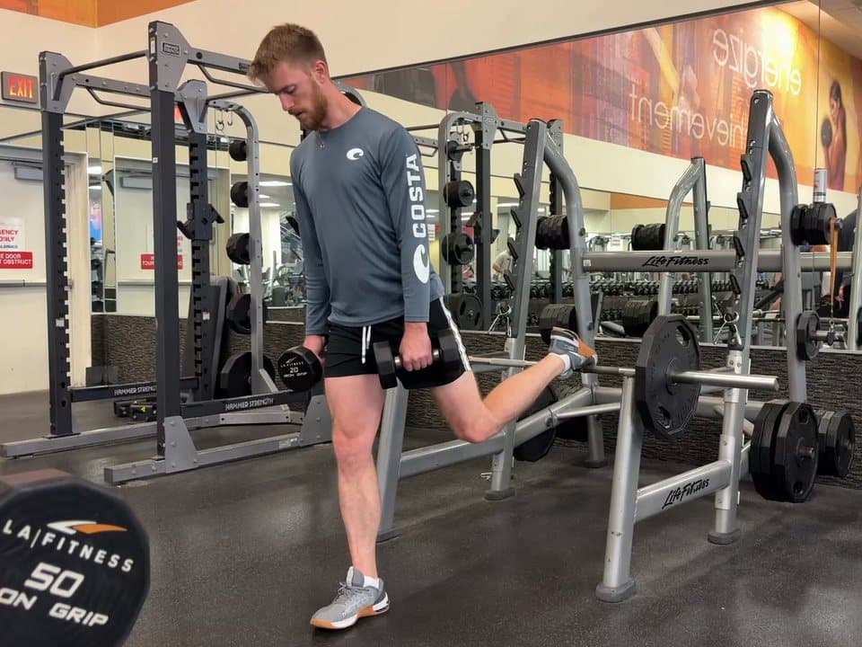 Connor Barth doing a Bulgarian split squat with dumbbells, his back foot up on a bar behind him.